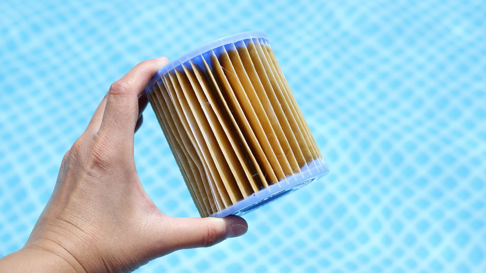 A hand holding a heavily stained pleated filter cartridge above pool water, the folds discoloured brown and grey from trapped organic buildup